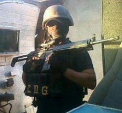 A man facing the camera and wearing a helmet, mask, and bullet proof vest holding a select-fire rifle.